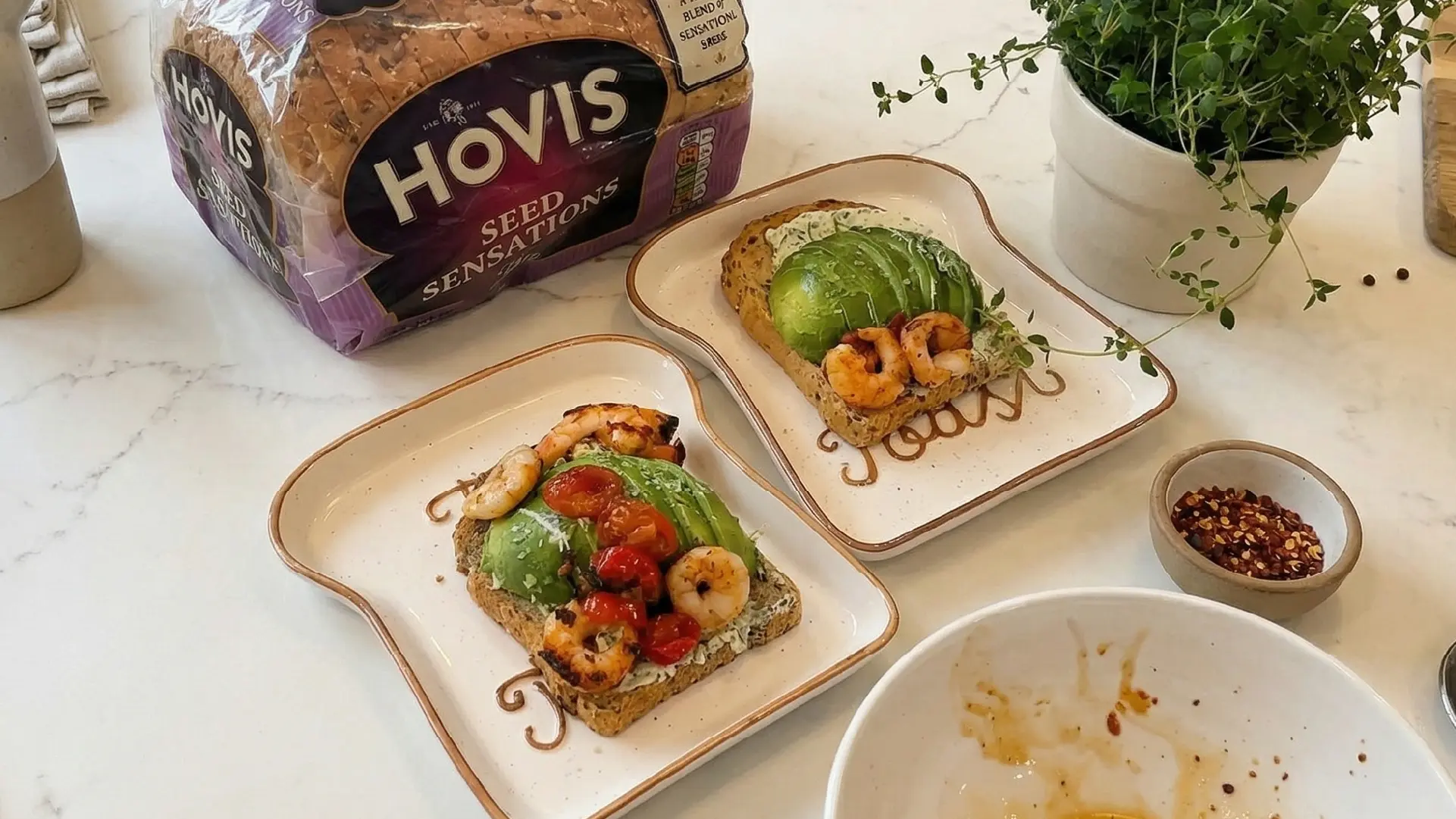 Assembling prawns and avocado toast on seeded bread, with Hovis loaf, chili flakes, and fresh herbs on a marble counter.