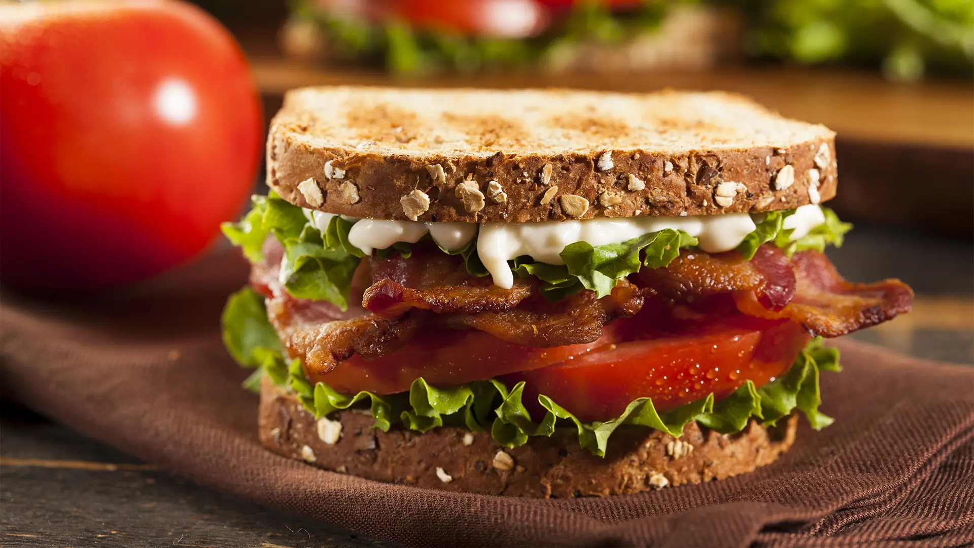 A classic BLT (bacon, lettuce, and tomato) sandwich sits on a brown cloth napkin. The sandwich is made with toasted whole wheat bread, crispy bacon, vibrant red tomato slices, fresh green lettuce, and a drizzle of white mayonnaise. A whole red tomato is visible in the blurred background to the left, and another partial sandwich and more lettuce are blurred in the background to the right.