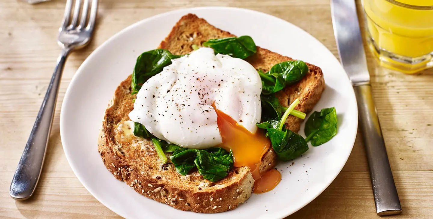 A slice of Hovis Granary sits on a plate with a poached egg and spinach on top. A knife and fork sits either side of the plate. 