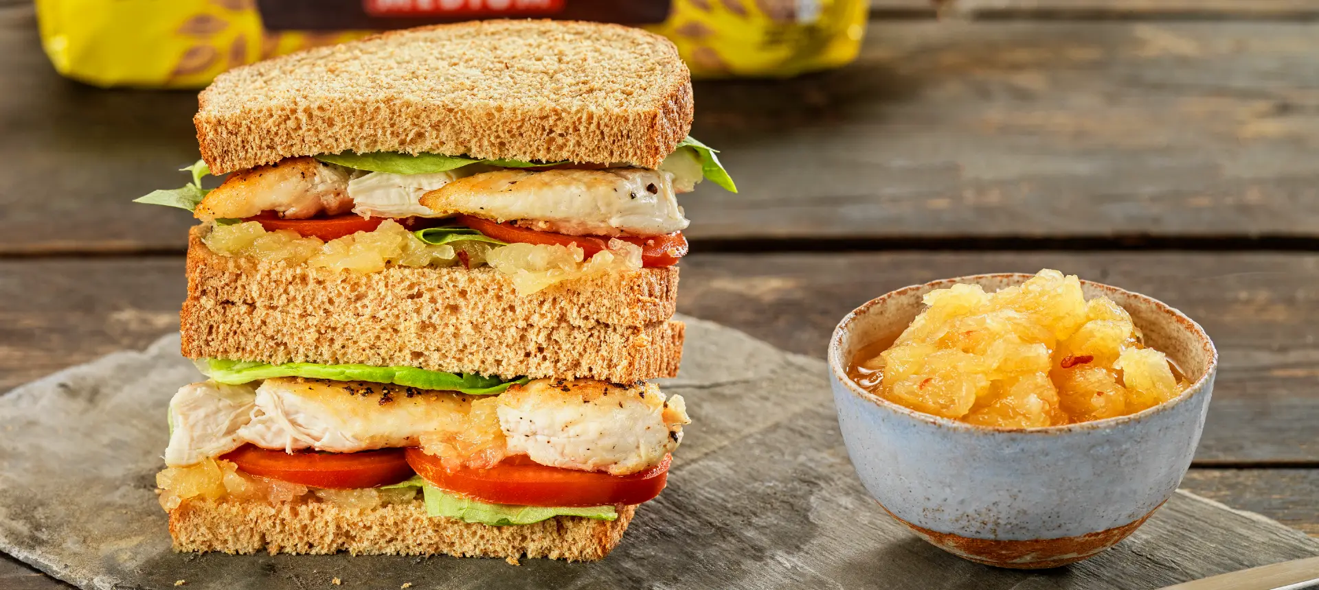 Two halves of a brown bread sandwich, stacked and filled with chicken, pineapple jam, and lettuce leaves, served with a small bowl of pineapple jam on the side.
