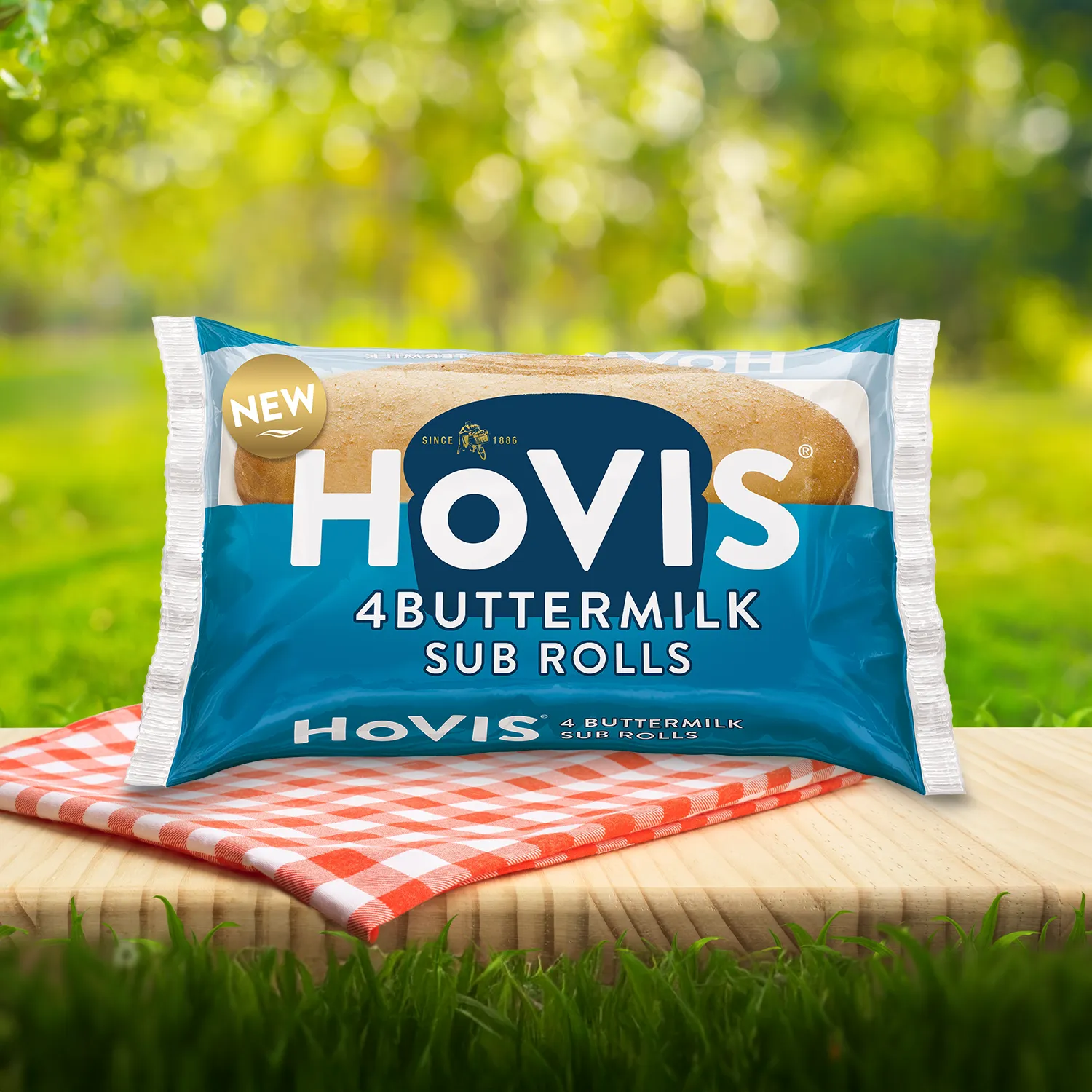 Hovis Buttermilk Sub Rolls packaging on a picnic table with a green outdoor background. The pack shows soft sub rolls  and highlights “Deliciously soft and flavourful with a hint of buttermilk.” A red gingham cloth adds a picnic feel to the setting.