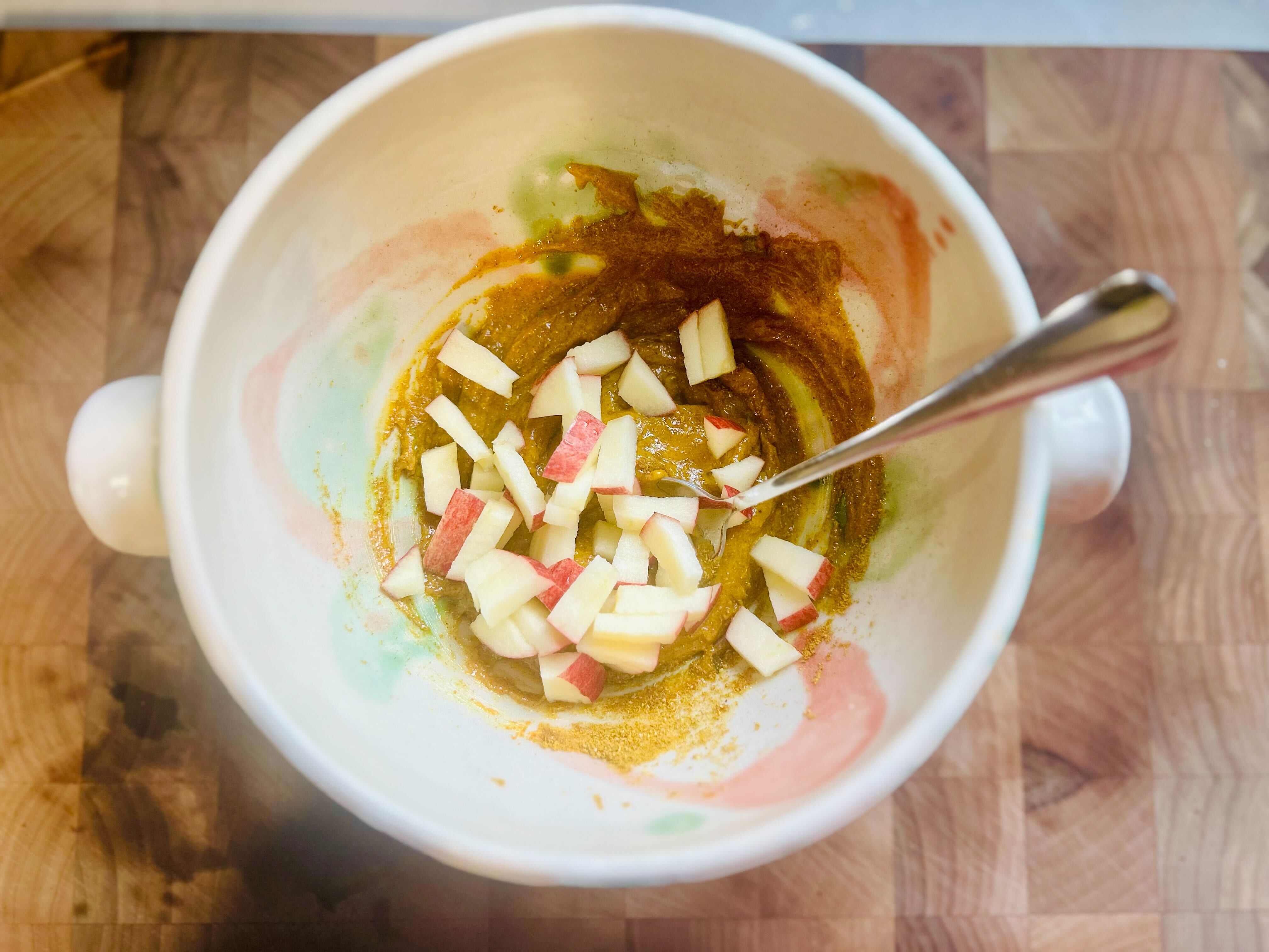 A close-up, top-down shot of a handmade ceramic bowl on a wooden chopping board. A metal spoon is stirring together a yellow curry-based sauce with finely diced apple chunks, some of which still show their red skin. The sauce appears to have a thick, creamy consistency.