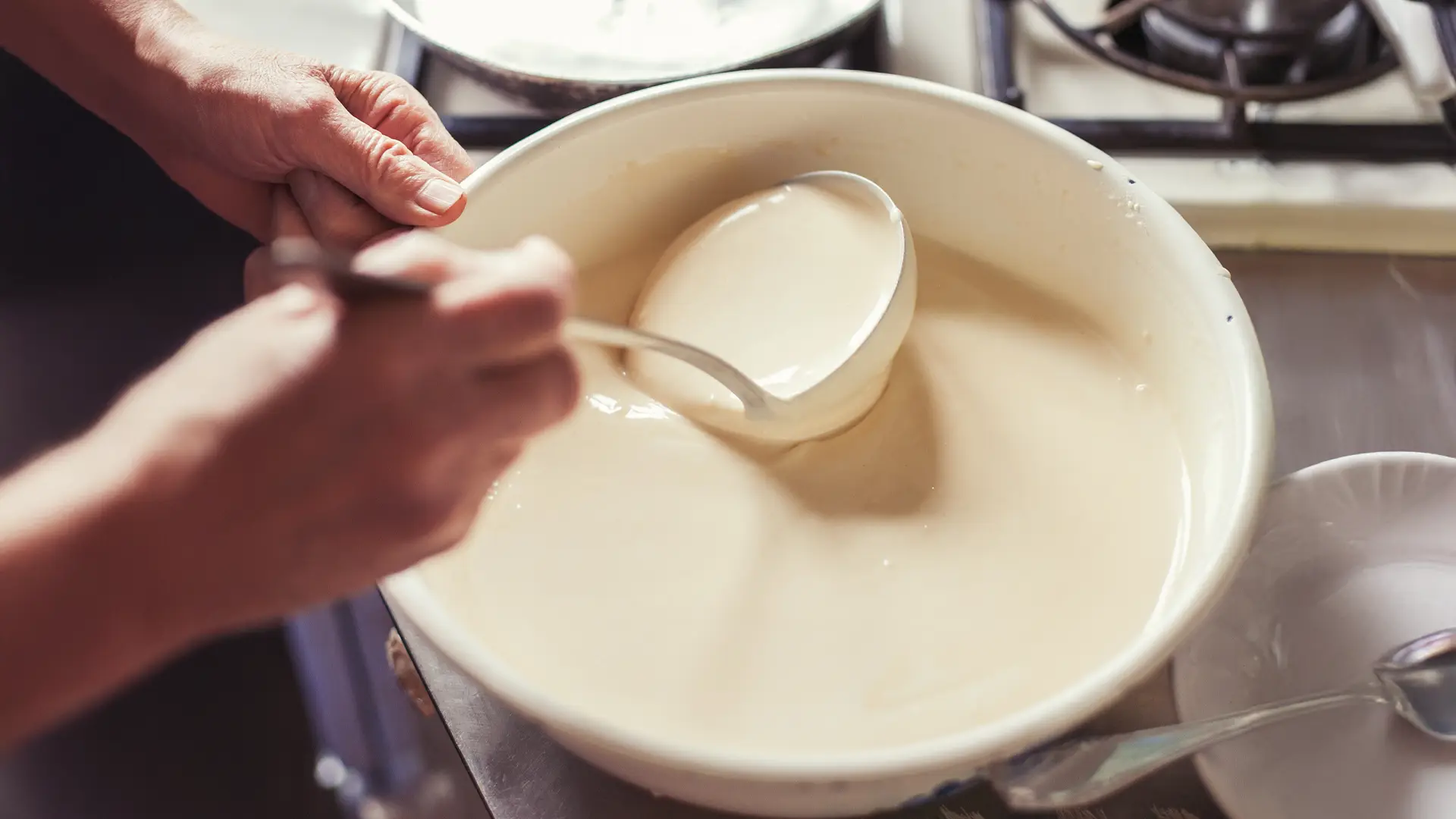 Someone uses a metal ladle to mix a smooth pancake batter in a medium bowl, ready to pour into a frying pan, on top of a hob.