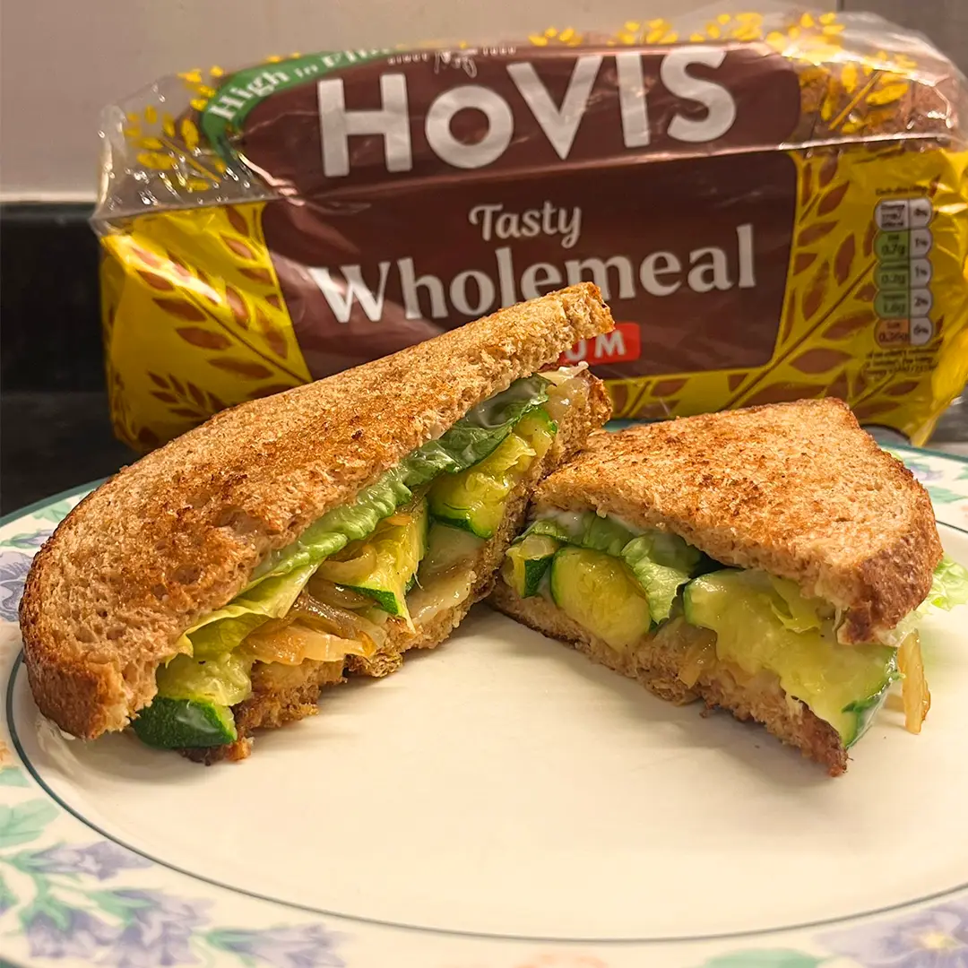 A toasted wholemeal sandwich with lettuce and courgette, cut in half and held by two hands. A loaf of wholemeal bread is in the background.
