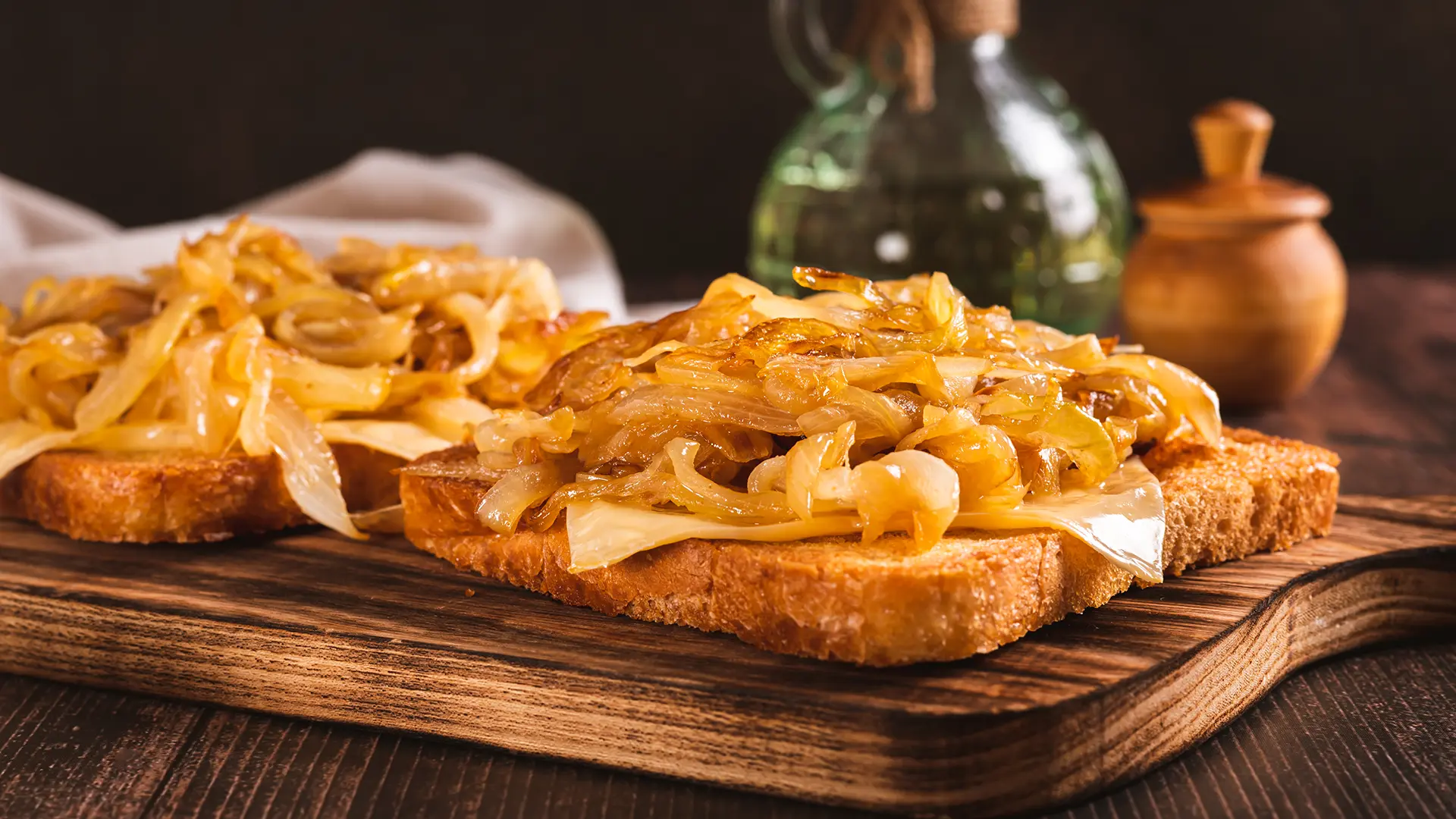 A close-up of two slices of toasted bread on a wooden chopping board, each topped with melted cheese and a serving of golden caramelised onions. In the blurred background, a glass bottle and a small wooden container are visible.