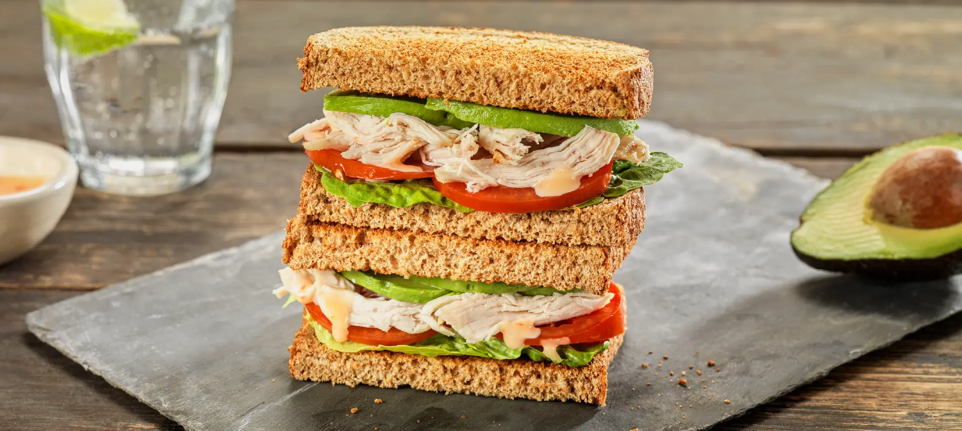 Two halves of a chicken sandwich made with Hovis Tasty Wholemeal Thick Sliced bread, filled with sliced avocado, tomatoes, and lettuce leaves.