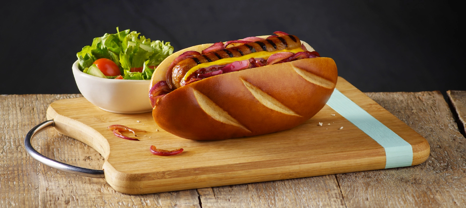 A hot dog made with Hovis Hot Dog Rolls, filled with a sausage, pickled red onions, and mustard, served with a side of green salad and tomatoes.