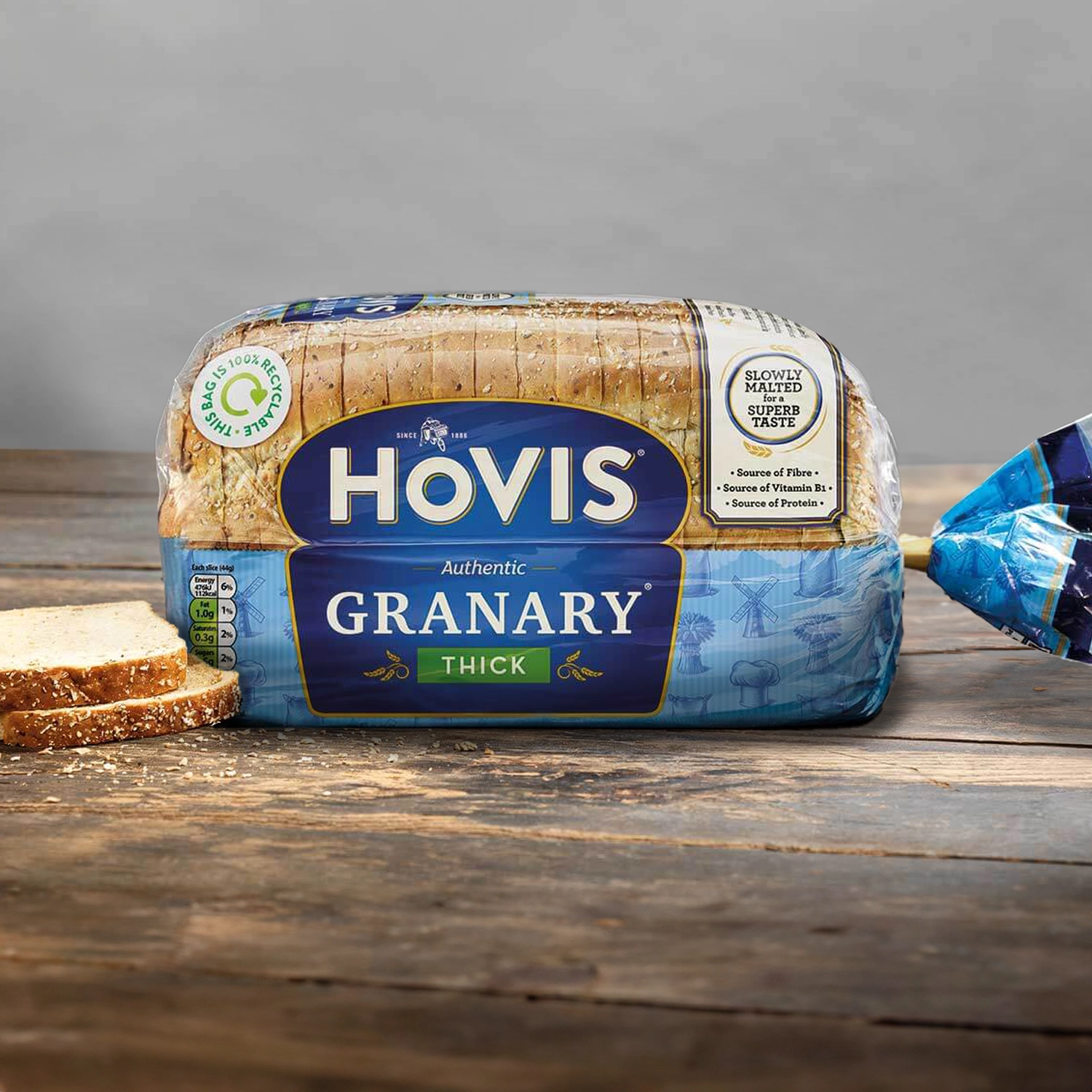 Hovis Granary Thick Sliced loaf against a light grey background, resting on a rustic wooden table with natural light, with two stacked slices of bread on the left.