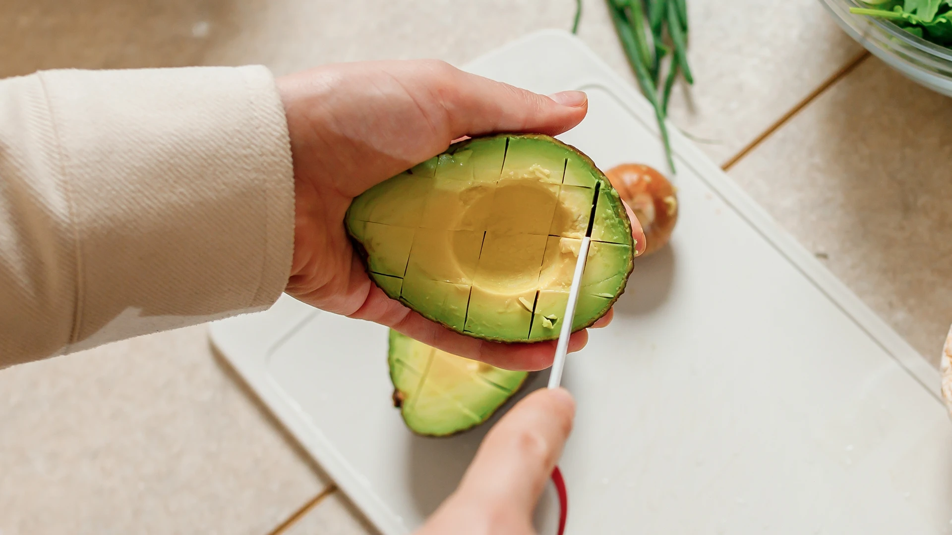 One hand holds half an avocado while the other slices it into cubes with a sharp knife on a white cutting board, with greens in the background.