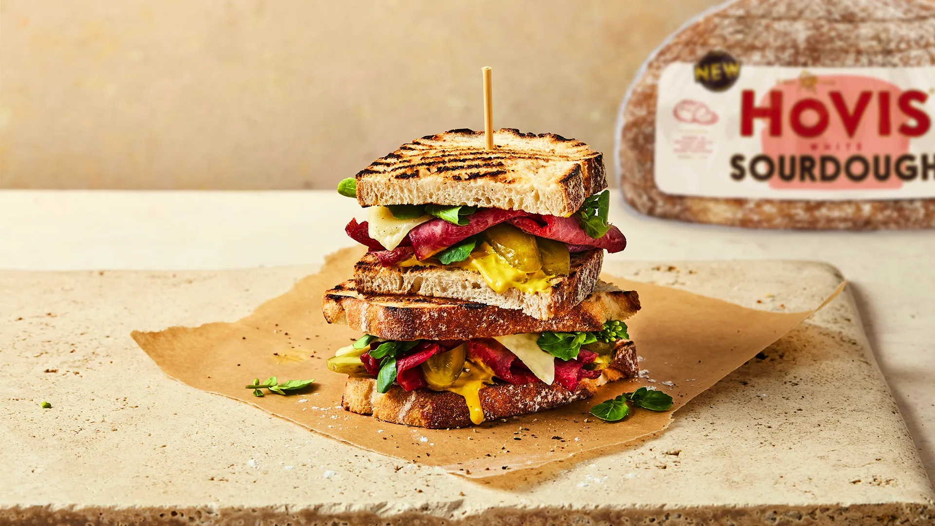 Two halves of a Hovis White Sourdough sandwich filled with cheese, pickles, spinach, and beef strips on a stone chopping board with the Hovis White Sourdough Loaf in the background