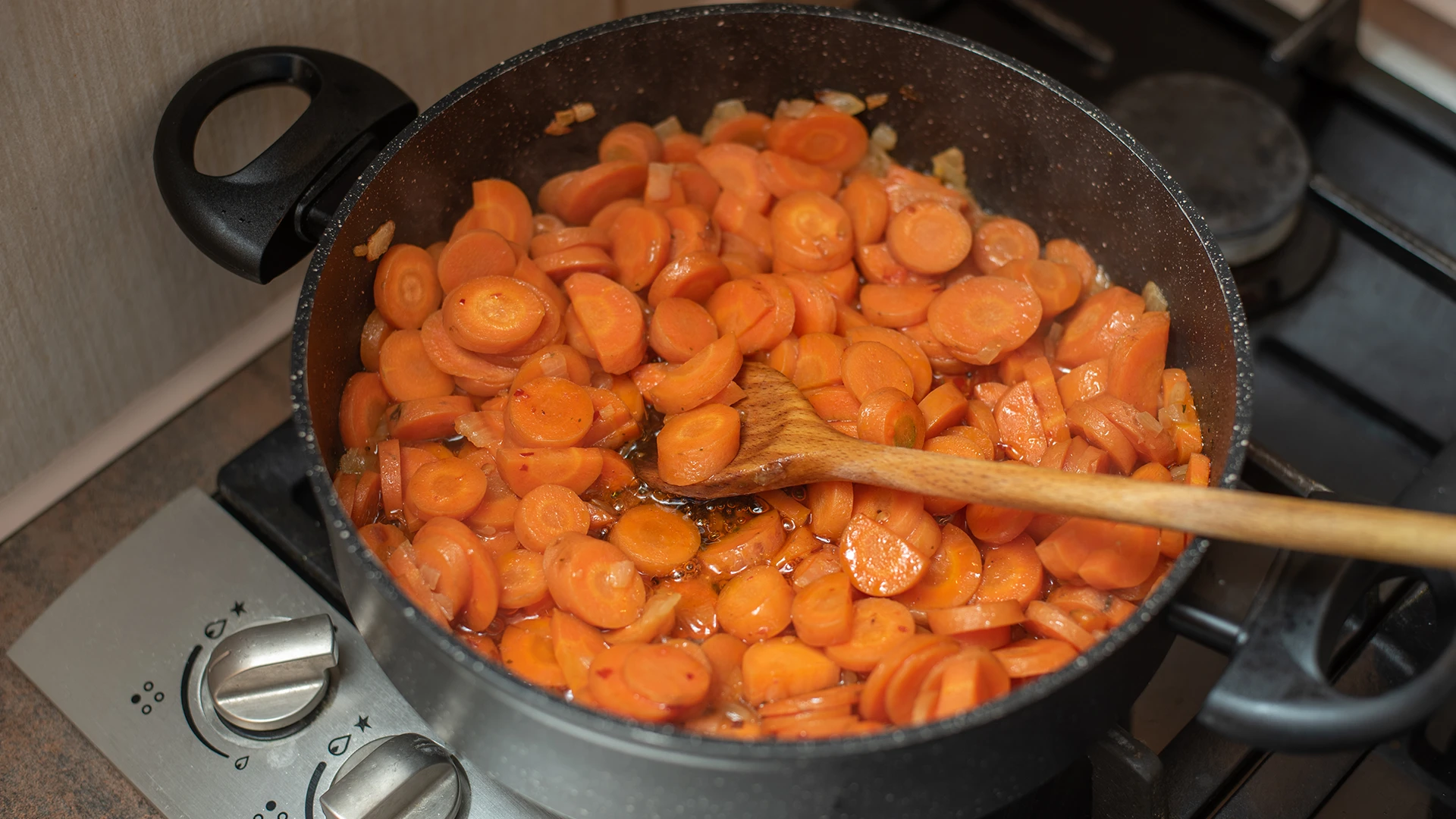 A large black pot on the hob contains round carrot slices, chopped onions, and liquid stock, with a wooden spoon resting inside.
