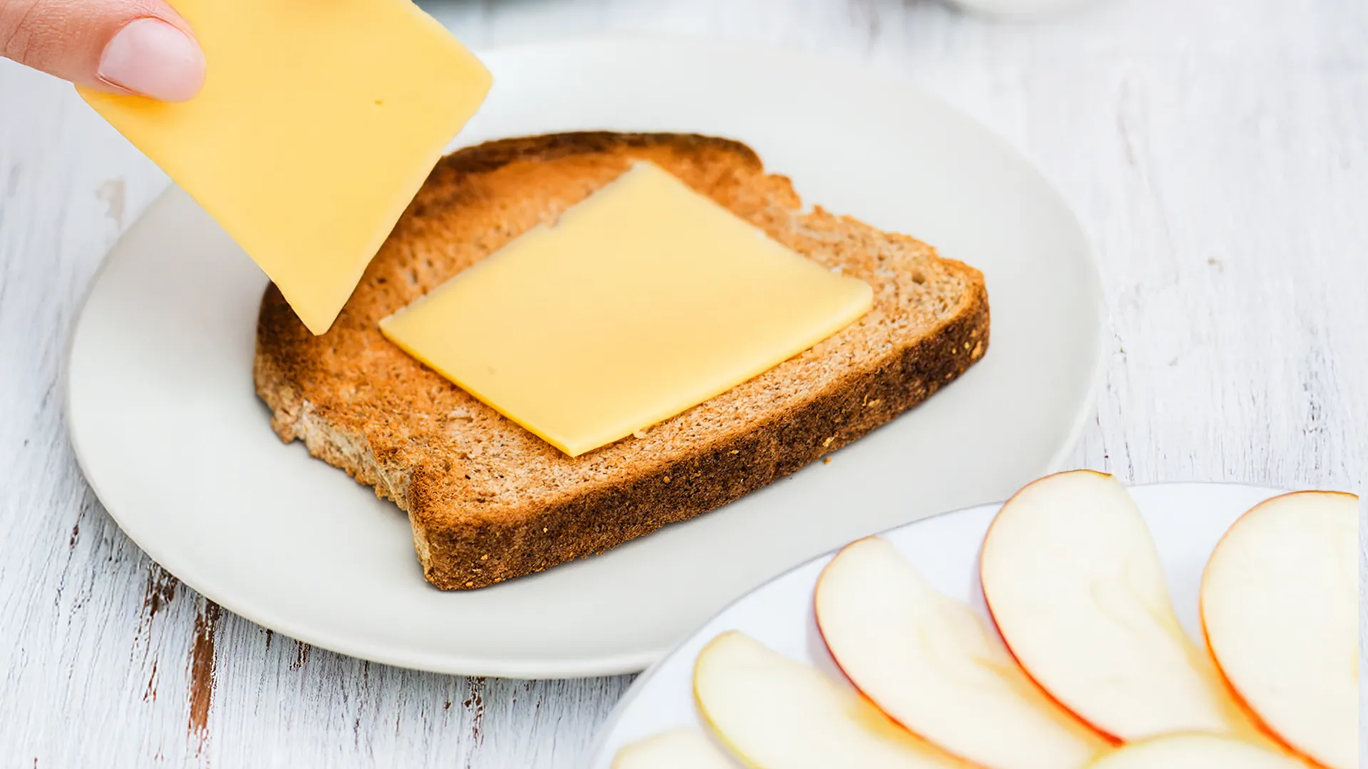 A hand places slices of cheddar on a toasted piece of Hovis® Farmhouse Wholemeal Batch with oat bread next to a plate of sliced apple pieces