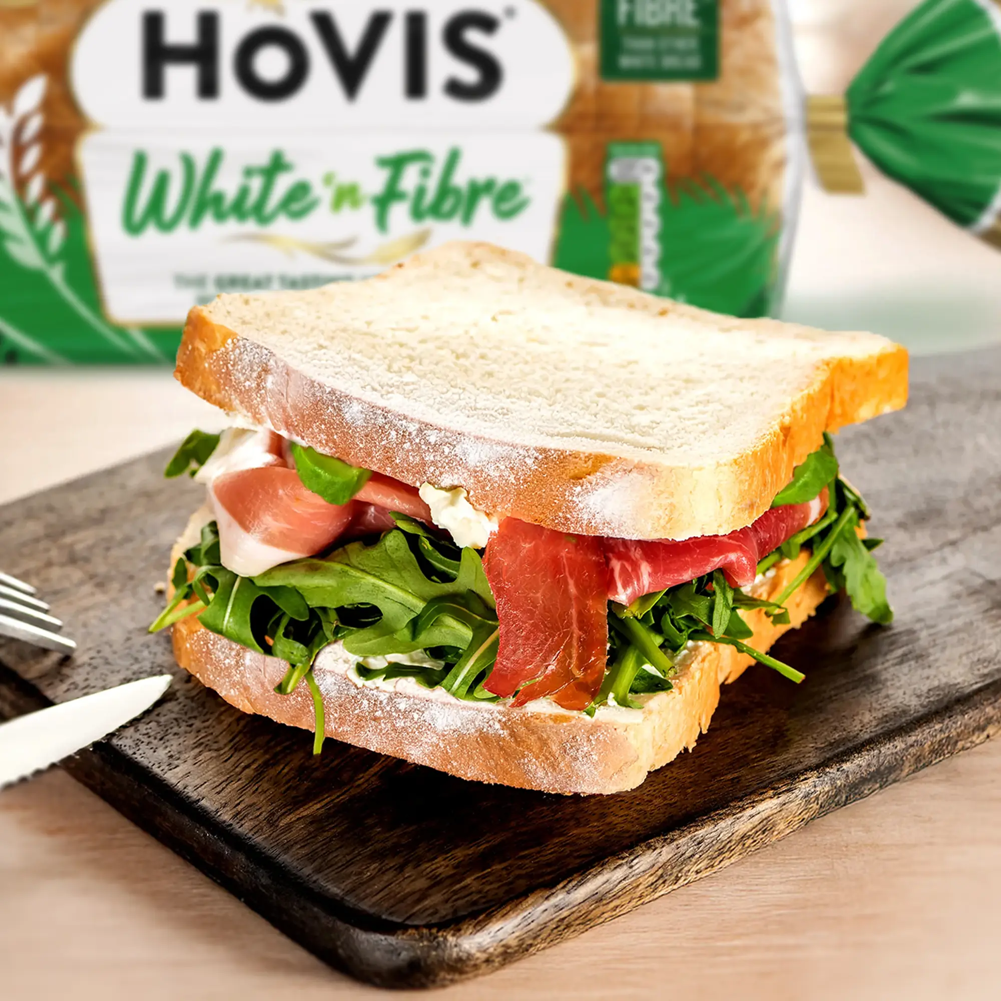 A sandwich made with Hovis White 'n Fibre bread sits on a dark wooden cutting board. The sandwich is filled with cheese spread, fresh rocket, and delicate slices of cured ham. The top slice of bread is slightly askew, revealing the delicious layers inside. A small portion of a knife and fork are visible to the left. In the background, a blurred loaf of Hovis White 'n Fibre bread in its green packaging adds context.