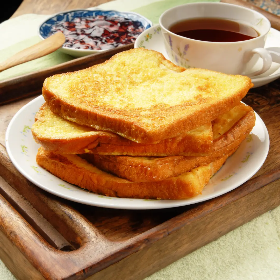 Four slices of toasted Hovis Soft White Extra Thick Sliced bread stacked on a plate, with a cup of tea and a small plate of jam on the side.