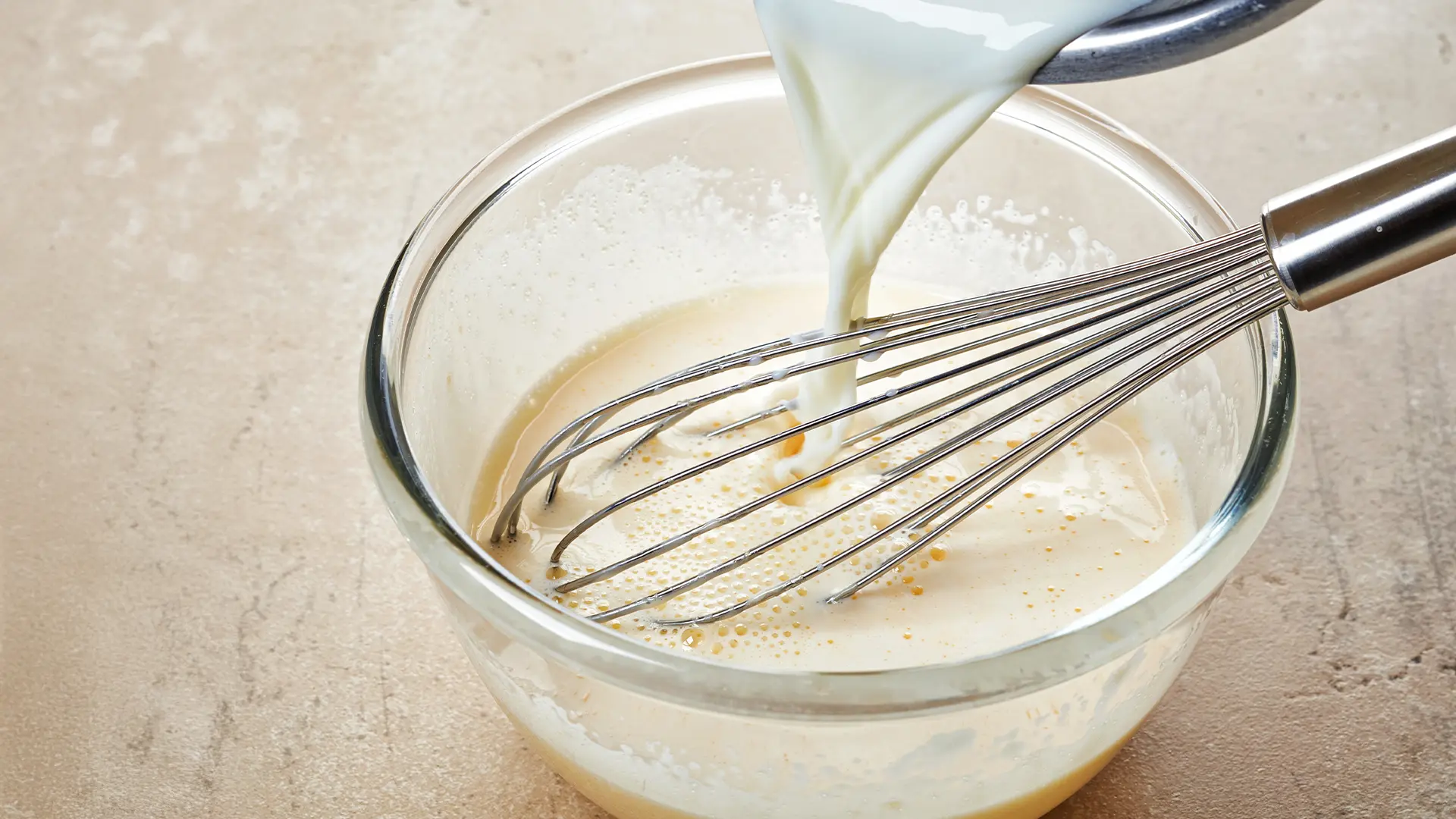 Someone pours in milk and uses a whisk to mix milk, egg and vanilla in a small, glass bowl, on top of beige, rough surface.