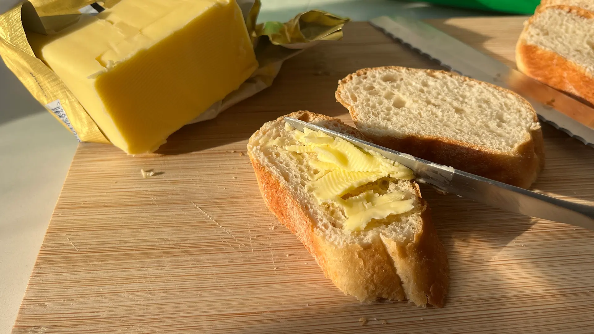 A close-up shot of a person using a silver butter knife to spread a thin layer of butter on a freshly cut slice of bread. The slice has a golden crust and a soft white interior. In the background, a block of butter is partially unwrapped in its foil, and another slice of bread and a serrated knife are visible on the wooden chopping board.