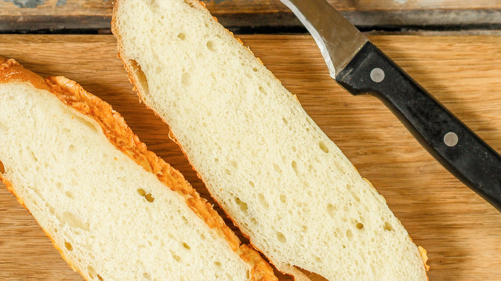 A cheese and herb sub roll cut in half lengthways on a chopping board next to a knife