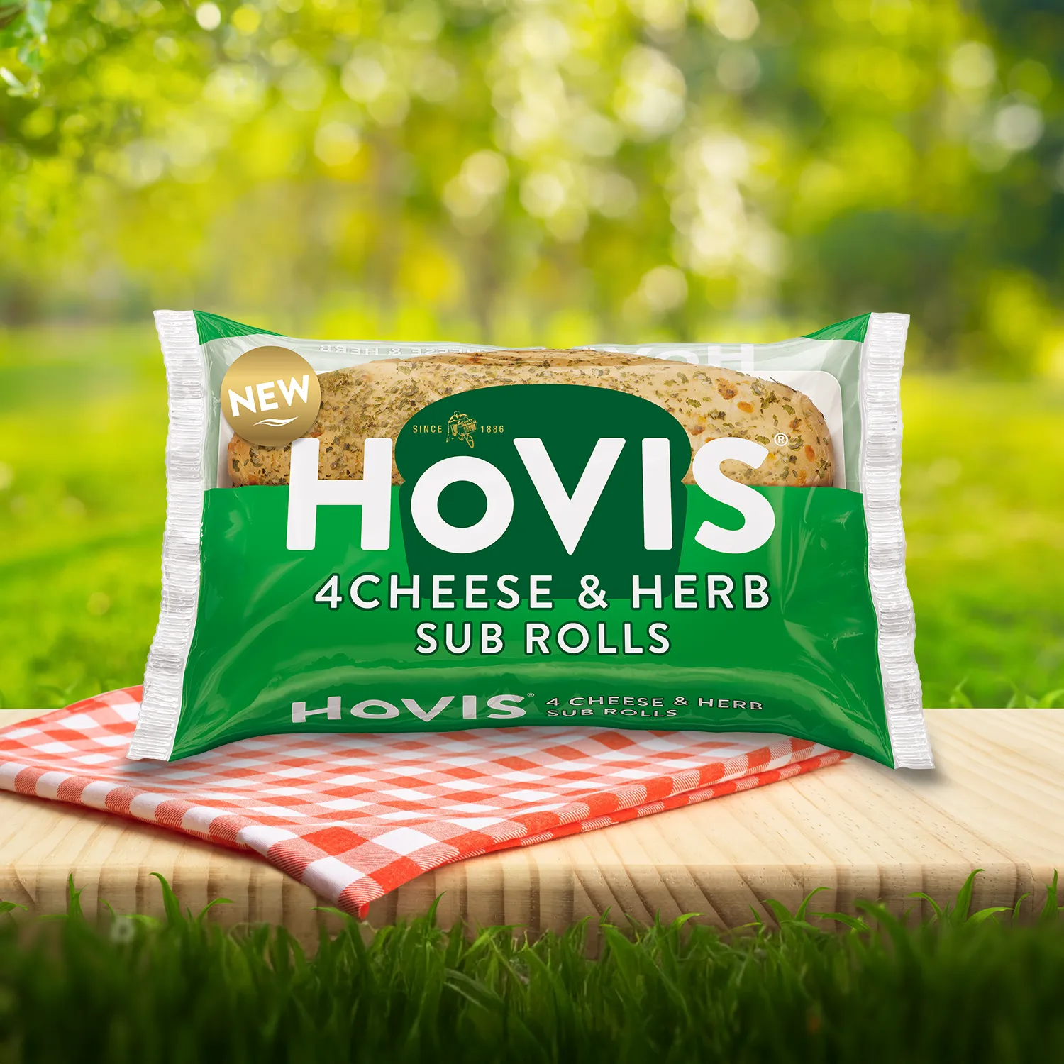Hovis Cheese & Herb Sub Rolls Sub Rolls packaging on a picnic table with a green outdoor background. The pack shows two soft sub rolls with herbs and highlights “Deliciously soft fusion of cheese and herbs.”  A red gingham cloth adds a picnic feel to the setting.