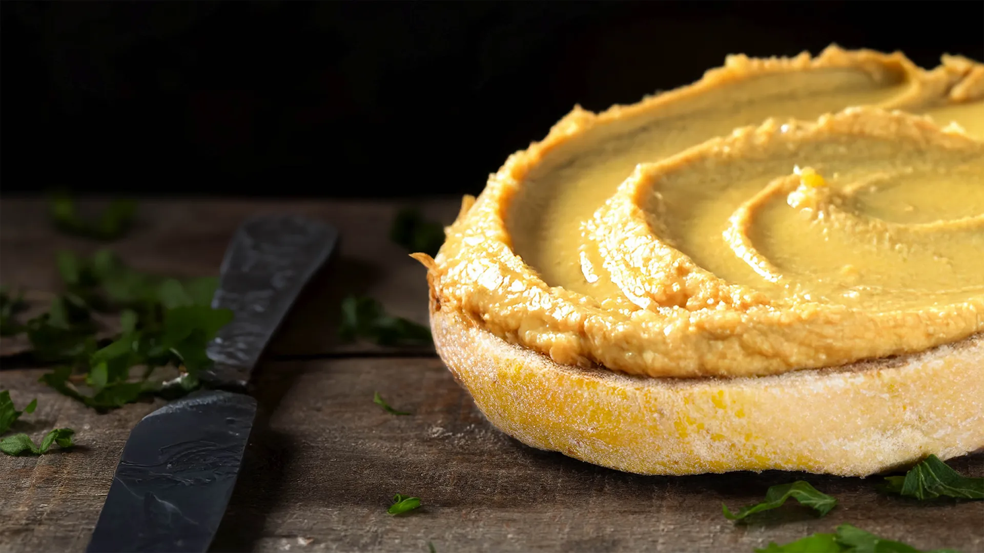 The bottom half of a sunflower and honey sub roll on a kitchen counter with thick hummus spread on top next to a spreading knife