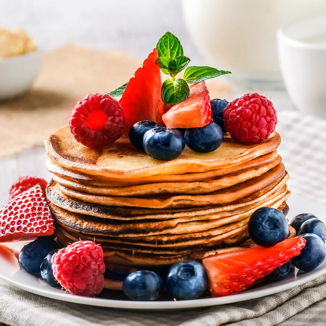 A stack of delicious bread pancakes rests on a white plate. They are topped with blueberries, raspberries, strawberries and mint. In the background a small bowl of almonds.