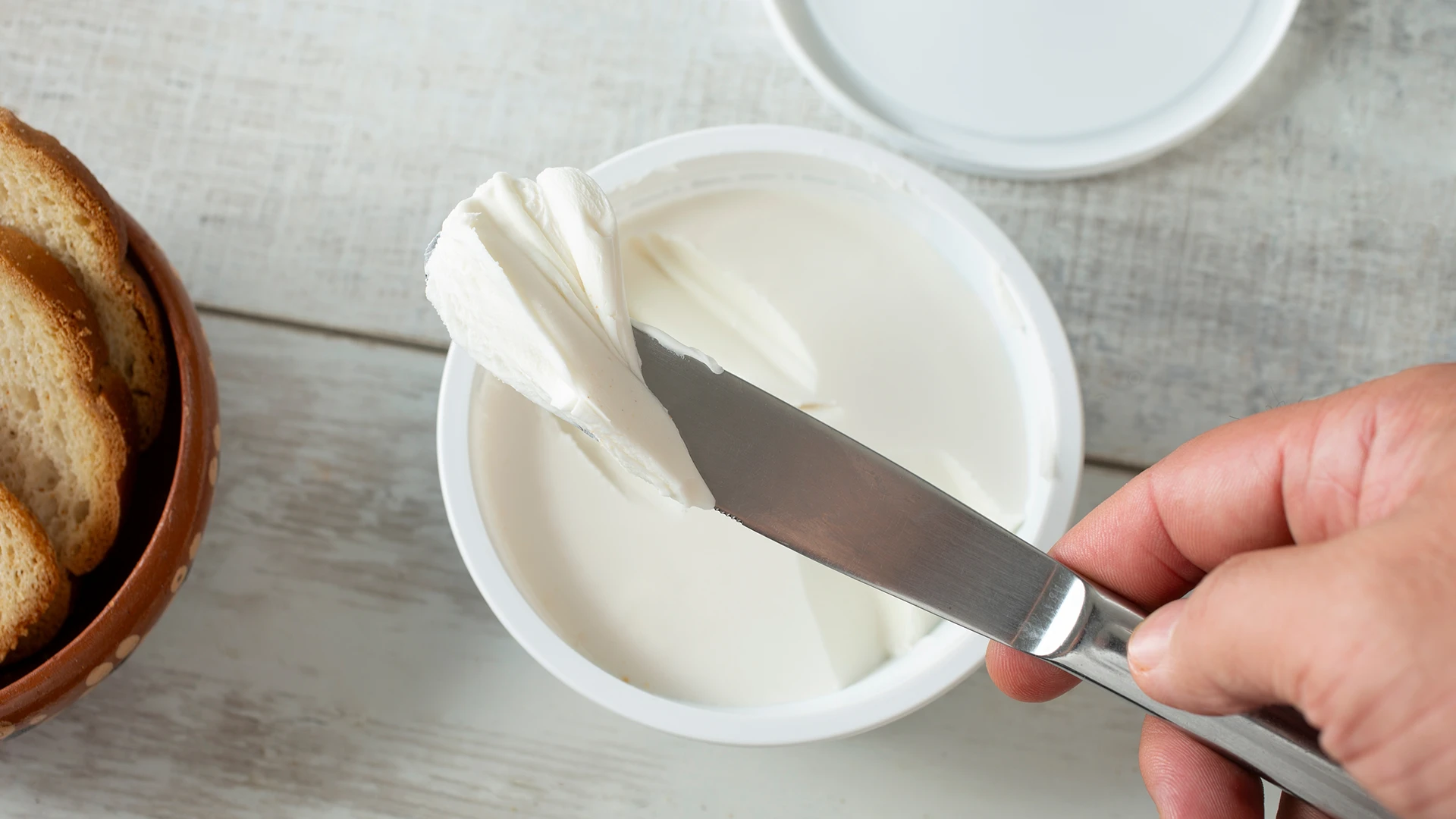 A hand holds a knife to scoop creamy white cream cheese from a plastic pot on a wooden table, with a brown bowl containing extra slices of bread.
