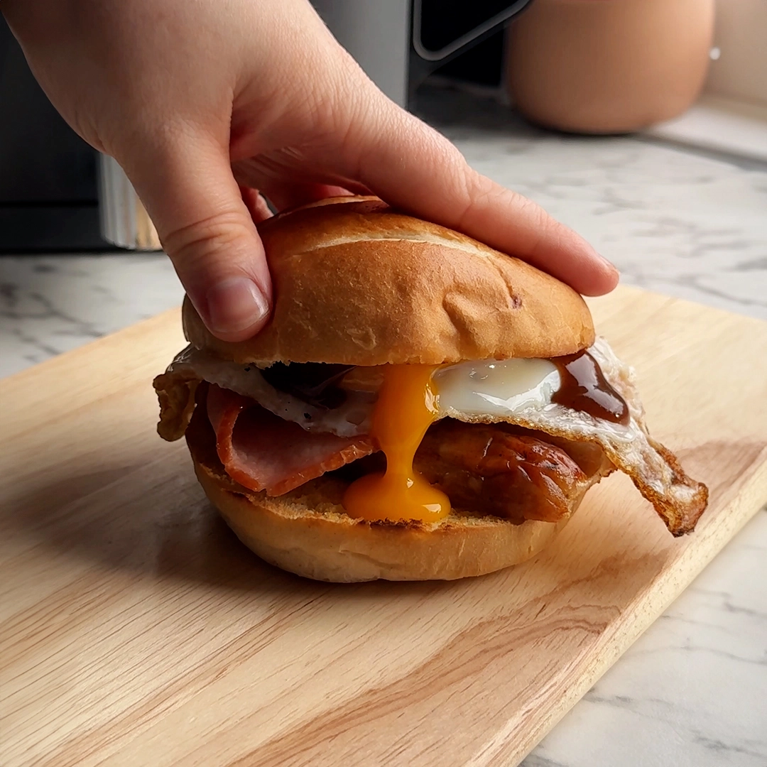 A hand holds a breakfast bun made with Hovis Burger Buns, filled with crispy bacon, sausage, and an egg with golden yolk oozing from it.