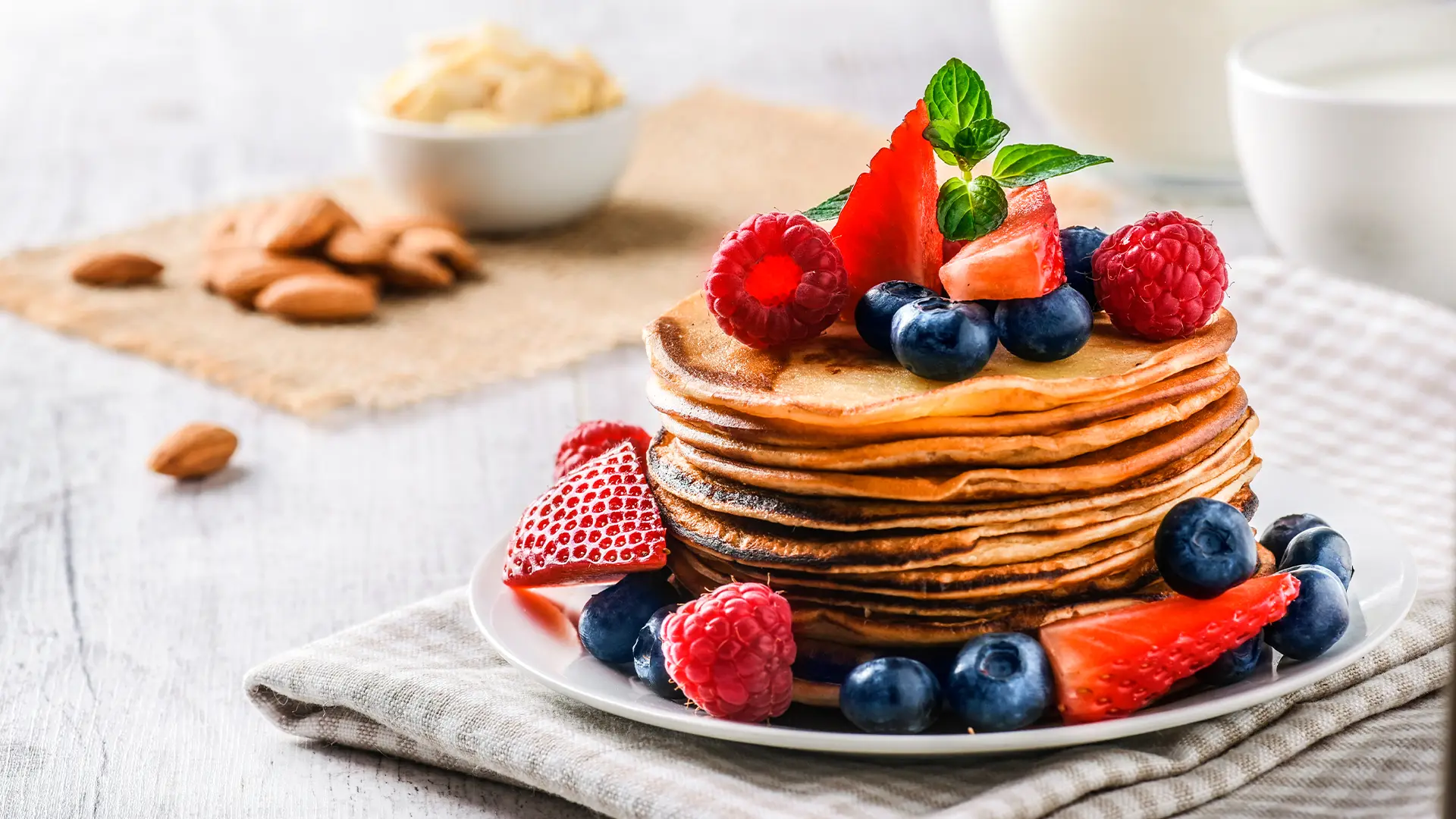 A stack of delicious bread pancakes rests on a white plate. They are topped with blueberries, raspberries, strawberries and mint. In the background a small bowl of almonds.